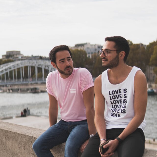 Zwei Personen, die an einem Fluss auf einer Mauer sitzen. Auf dem Oberteil einer Person steht “Love is”.